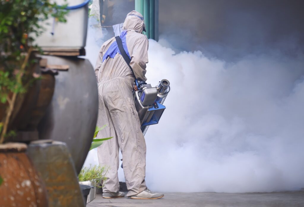 Rear view of outdoor healthcare worker using fogging machine spraying chemical to eliminate mosquitoes and prevent dengue fever on overgrown at slum area